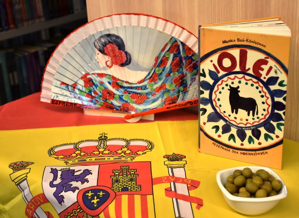Flaga Hiszpanii, w tle rozwinięty wachlarz, książka i miseczka z oliwkami